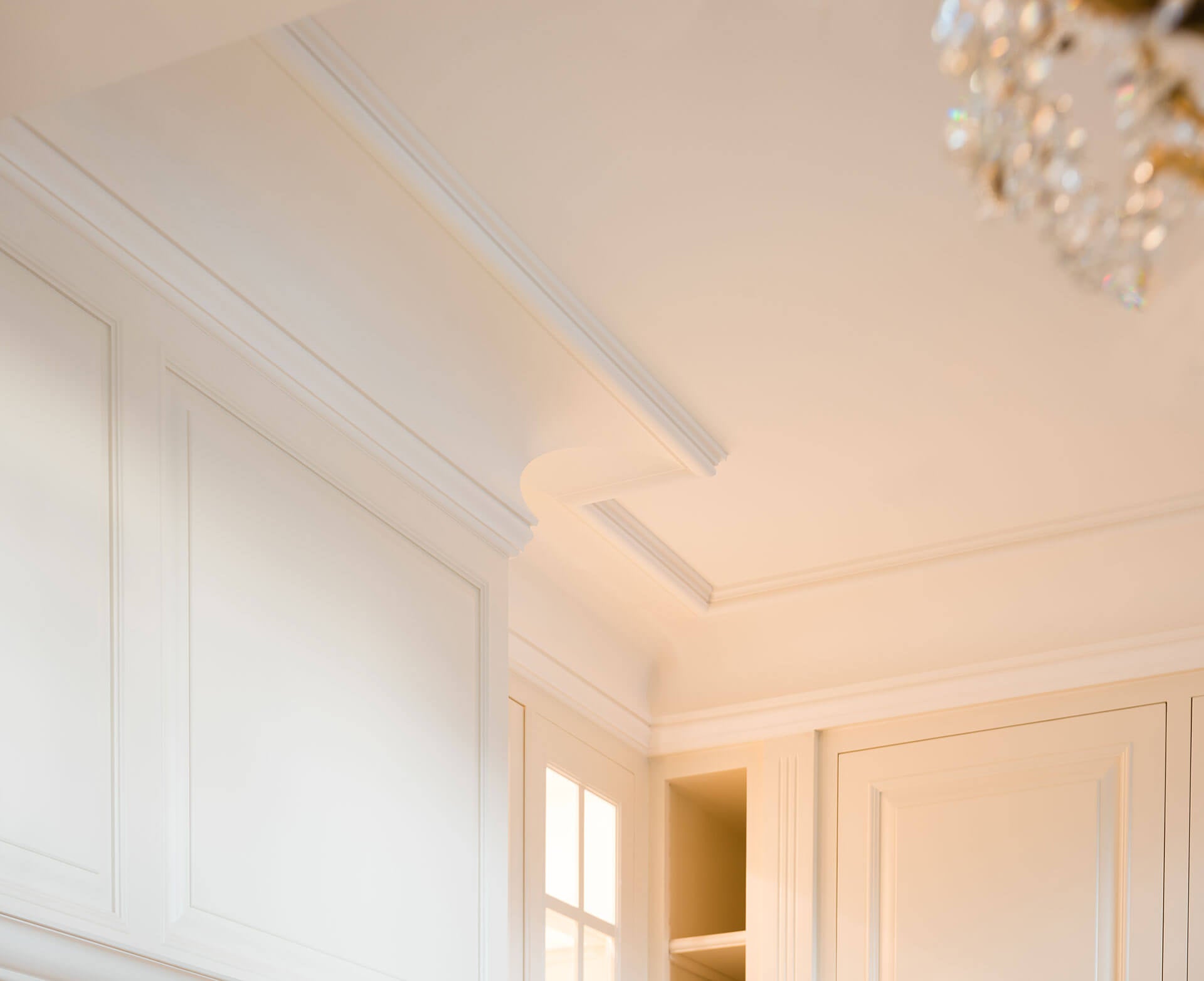 Eine Nahaufnahme einer eleganten, cremefarbenen Decke mit der HERITAGE XL Stuckleiste von Orac Decor strahlt klassische Eleganz aus. Ein Blick auf einen Kronleuchter schmückt die obere rechte Ecke, während subtiles natürliches Licht durch ein Fenster oder eine Tür fällt und die zeitlose Purotouch-Eleganz widerspiegelt.