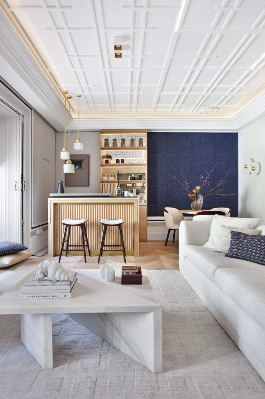 Ein modernes Wohnzimmer verfügt über eine geometrisch gemusterte Decke, die mit der C327 MANOIR Stuckleiste von Orac Decor dekoriert ist, und einen Parkettboden. Es präsentiert ein weißes Sofa, einen Marmor-Couchtisch, einen Holzbarbereich mit Hockern und einen Esstisch, der von Stühlen umgeben ist. Eine strukturierte blaue Akzentwand sorgt für einen auffälligen Kontrast.