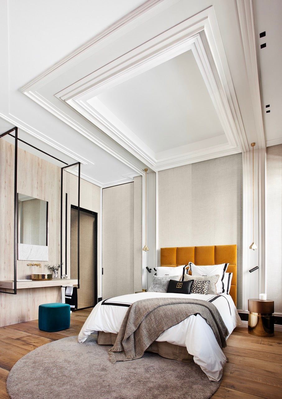 Ein modernes Schlafzimmer verfügt über ein großes Bett mit weißer und grauer Bettwäsche und einem orangefarbenen Kopfteil. Das Zimmer verfügt über Holzböden, einen gemütlichen runden Teppich und einen eingebauten Schminktisch mit Spiegel. Hohe Wände werden durch die Orac Decor C360 Stuckleiste aufgewertet, während die Kassettendecke aus Purotouch-Material das stilvolle Dekor vervollständigt.