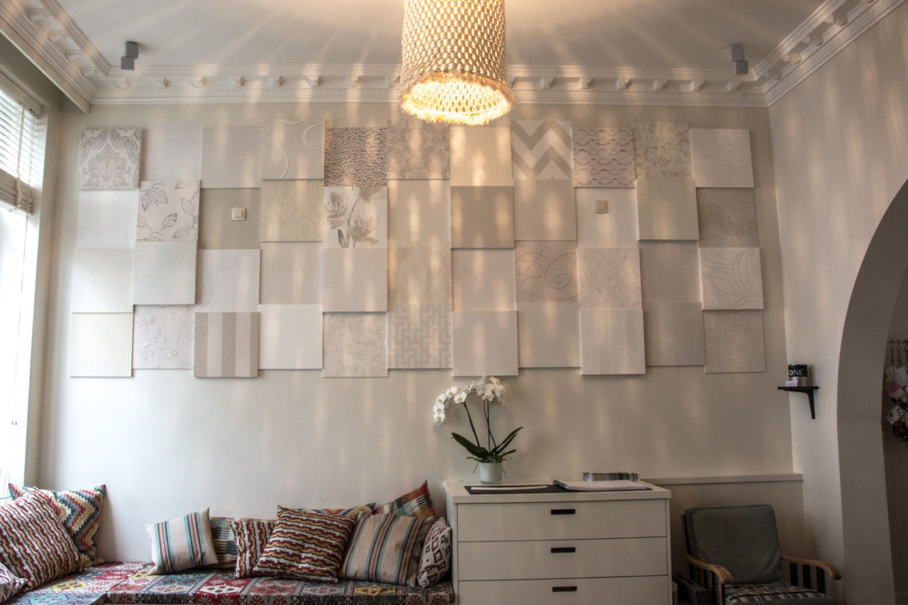 Eine Wand mit einem dekorativen Arrangement aus weißen und hellgrauen quadratischen Orac Decor Duropolymer-Paneelen, beleuchtet von einem Kronleuchter. Darunter steht ein modernes Sideboard, geschmückt mit einer weißen Orchideenvase. Auf einer Bank an der Wand liegen bunt gemusterte Kissen.