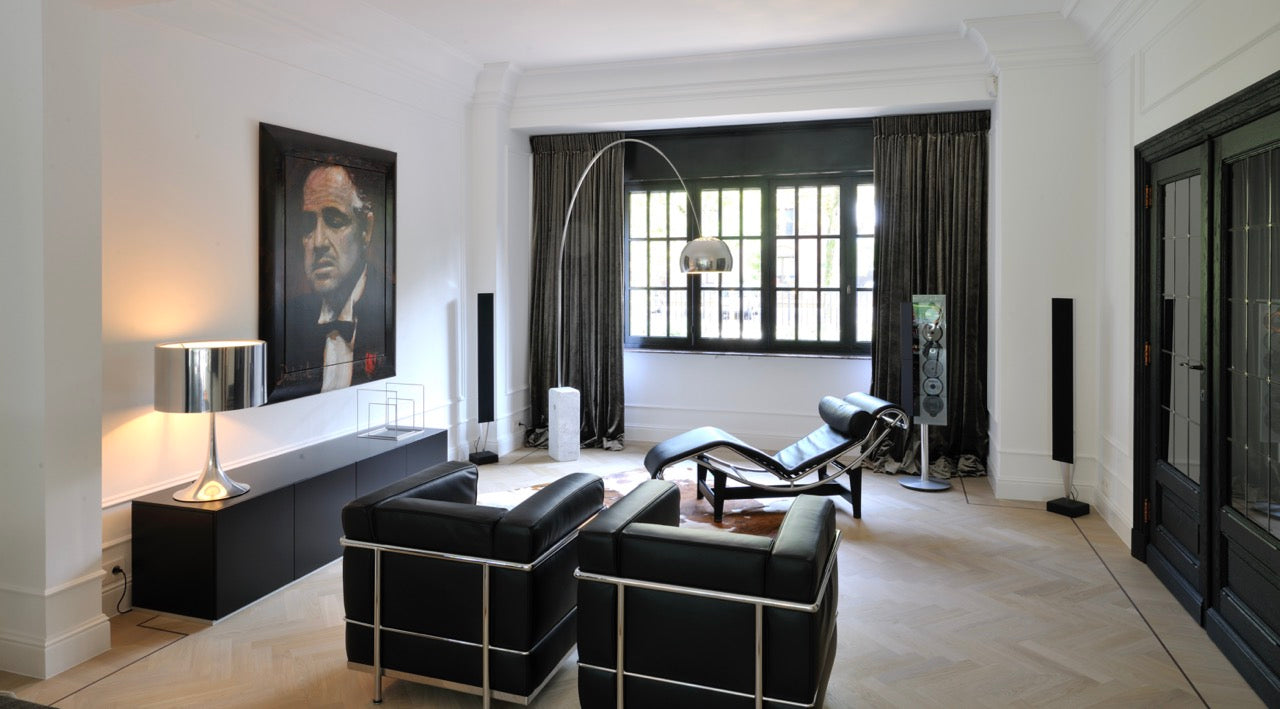 Ein modernes Wohnzimmer ist mit schwarzen Lederstühlen, einer stilvollen Chaiselongue und einer großen gewölbten Stehlampe ausgestattet. Ein großes gerahmtes Porträt wertet die mit Orac Decor-Elementen dekorierte weiße Wand auf, während dunkle Vorhänge das Fenster einrahmen. Ein Konsolentisch mit einer metallischen Lampe wertet das anspruchsvolle Dekor auf.