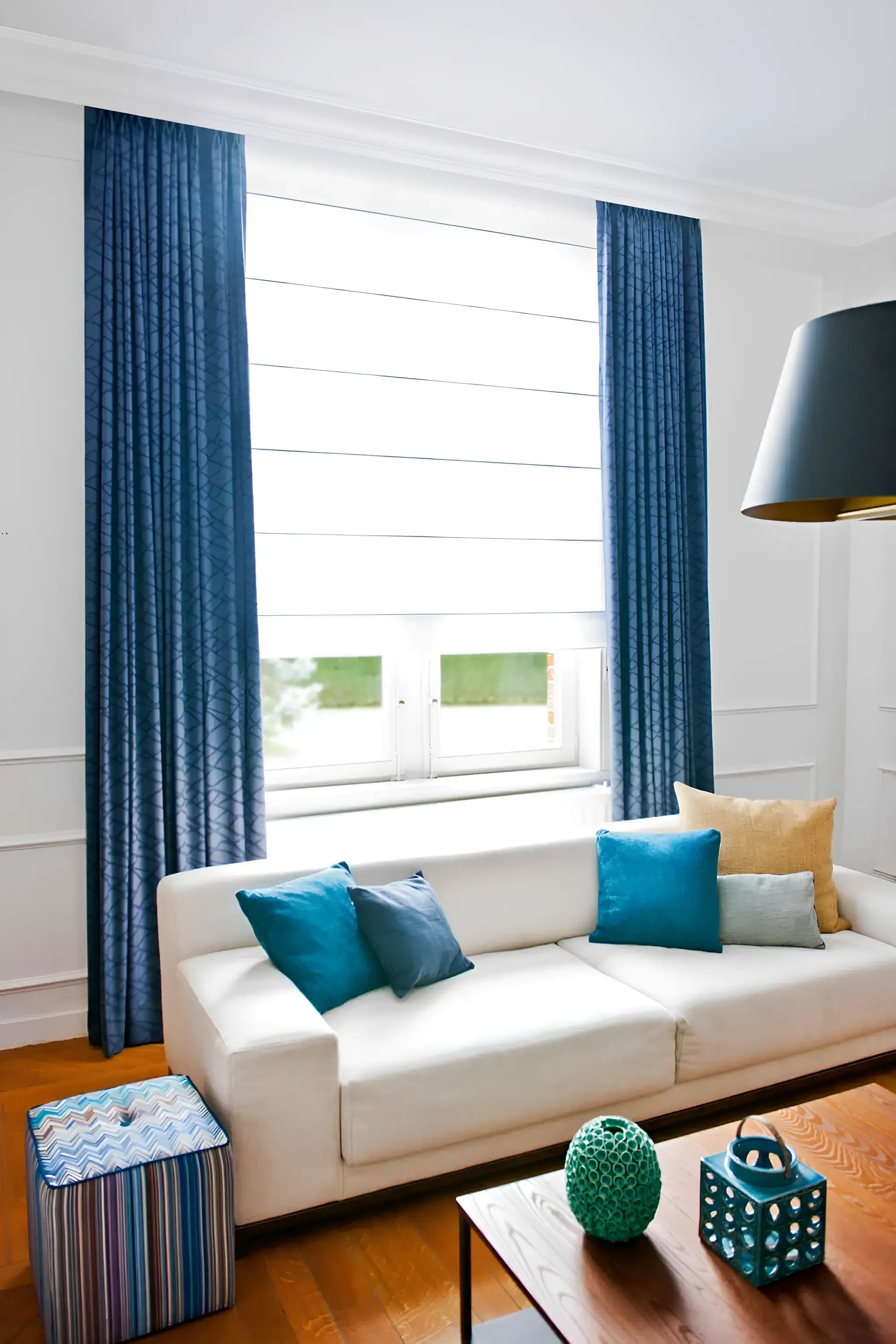 Ein modernes Wohnzimmer, das klassische Eleganz ausstrahlt, präsentiert ein weißes Sofa mit blauen und beigen Kissen. Blaue Vorhänge rahmen ein großes Fenster ein, das den Raum mit natürlichem Licht verschönert. Ein Ottoman mit geometrischem Muster und dekorative blaugrüne Gegenstände setzen stilvolle Akzente auf dem Holzboden, alles elegant hervorgehoben durch die HERITAGE M Stuckleiste von Orac Decor.