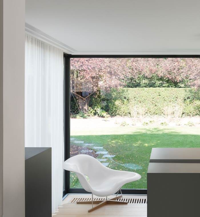 Ein moderner Raum verfügt über ein großes, vom Boden bis zur Decke reichendes Fenster mit Blick auf einen üppigen Garten. Neben dem Fenster steht ein stilvoller weißer Stuhl, eingerahmt von transparenten Vorhängen und aufgewertet durch die C391 STEPS-Stuckleiste von Orac Decor. Das natürliche Licht durchflutet das mit Purotouch durchtränkte Interieur und verkörpert moderne Raumkonzepte.