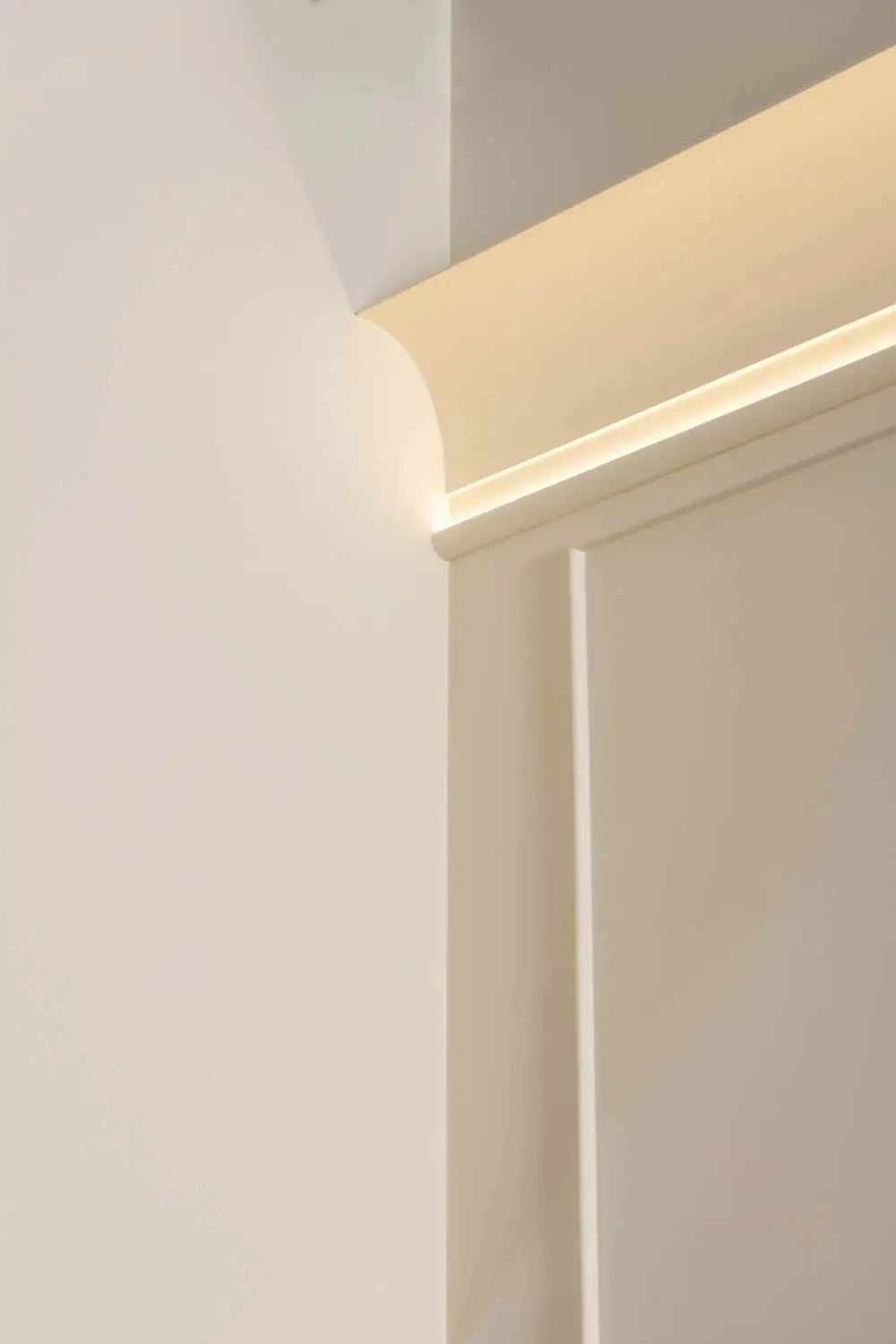 Nahaufnahme minimalistischer Innenarchitektur, die eine glatte, beige Wandecke mit abgerundeter Kante und sanfter Beleuchtung von oben hervorhebt, die sanfte Schatten wirft. Die Orac Decor C990 INFINITY Stuckleiste verleiht einen Hauch französischen Landhaus-Charme und schafft eine harmonische und elegante Atmosphäre.