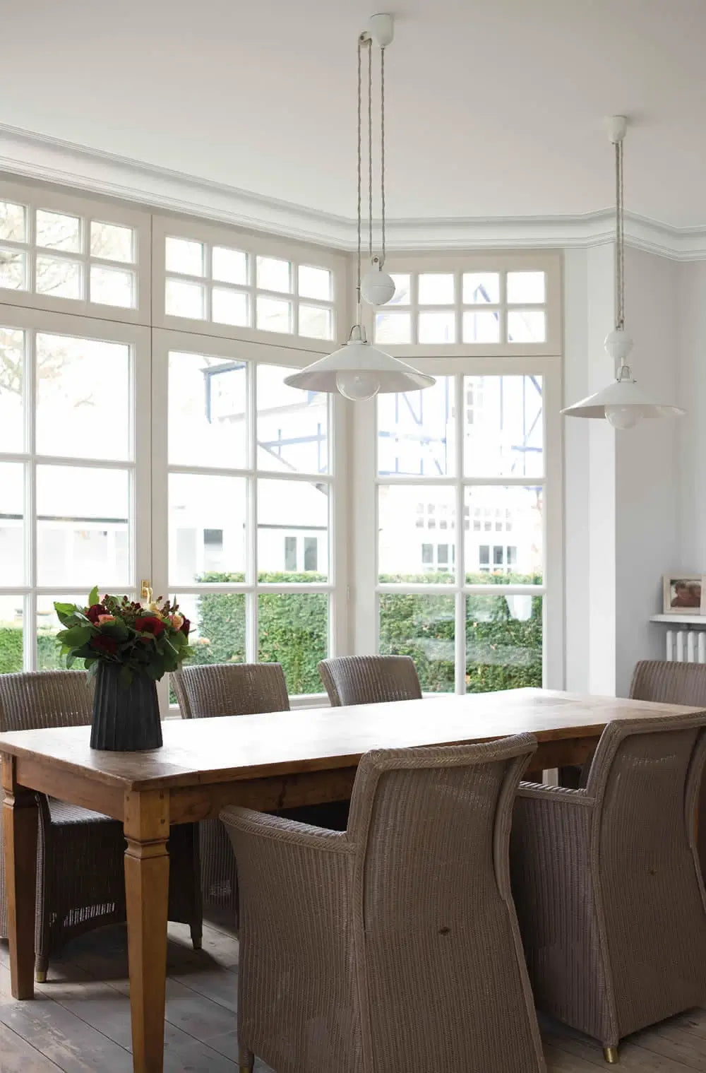 Ein helles Esszimmer im klassischen Stil verfügt über einen großen Holztisch, der von Korbstühlen umgeben ist. Darüber hängen zwei weiße Pendelleuchten aus Duropolymer von Orac Decor. Große Fenster mit Gittermuster bieten einen Blick ins Grüne draußen und eine Vase mit Blumen ziert den Tisch.