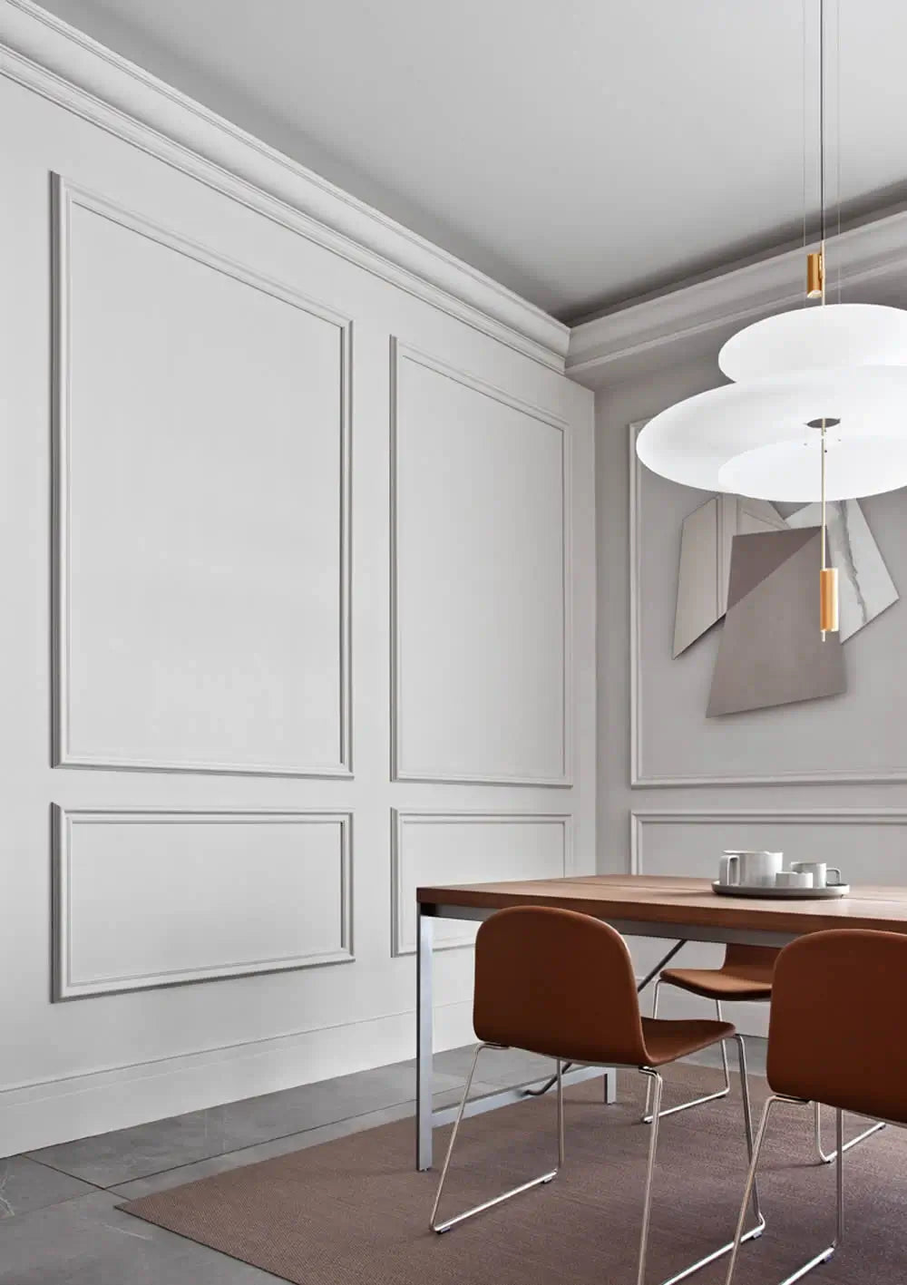 Ein minimalistisches Esszimmer präsentiert weiße Purotouch-Wände von Orac Decor, aufgewertet durch dekorative Zierleisten und eine Deckenleuchte. Ein Holztisch wird durch zwei braune Stühle ergänzt, die auf einem grauen Teppich ruhen. Darüber sorgt eine moderne, mehrschichtige weiße Pendelleuchte für Eleganz, während abstrakte Kunst die Wand für eine anspruchsvolle Note ziert.
