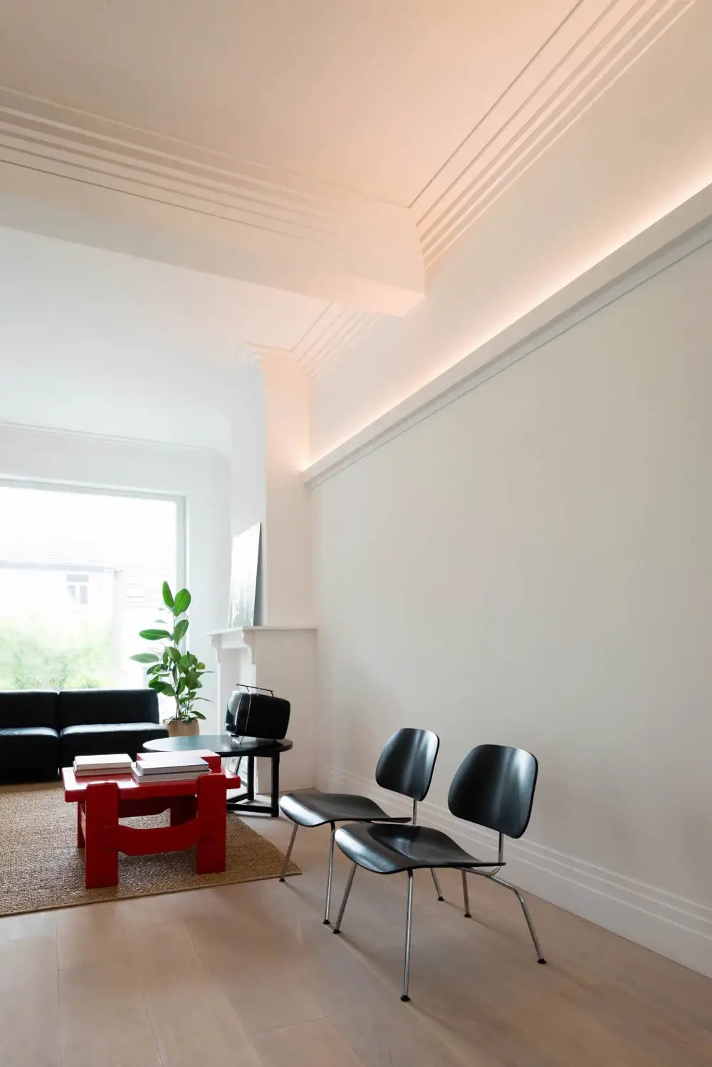 Das minimalistische Wohnzimmer ist mit zwei schwarzen Stühlen, einem modernen roten Couchtisch und einem eleganten schwarzen Sofa ausgestattet. Natürliches Licht fällt durch ein großes Fenster und betont die weißen Wände, die mit der dezenten Orac Decor C393 STEPS Stuckleiste geschmückt sind. Eine Topfpflanze und Bücher verleihen diesem modernen Profilraum Charme.