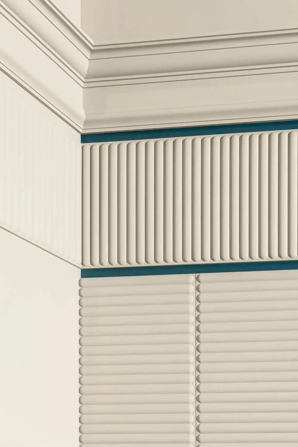 Eine Nahaufnahme einer beigen Wandecke zeigt dekorative vertikale Rippenpaneele mit blauen Akzenten, elegant eingerahmt von der C332 NOBLESSE-Kronenleiste von Orac Decor. Das Licht erzeugt sanfte Schatten und betont die Texturen und architektonischen Elemente in diesem Purotouch-Design.